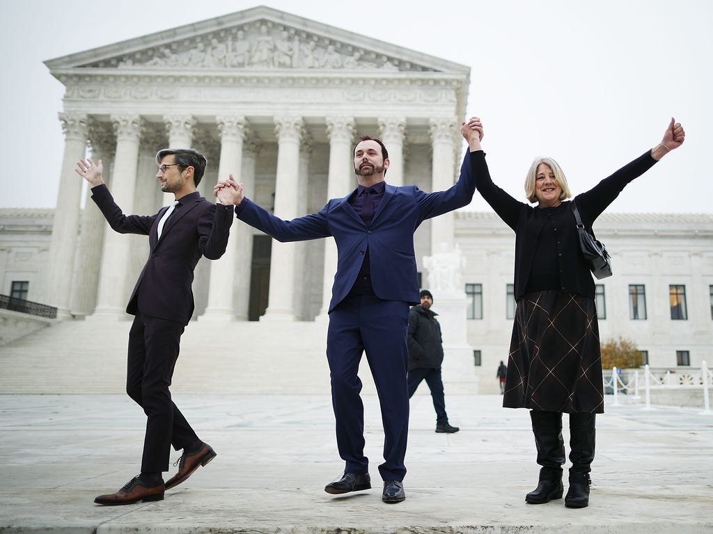 (L-R) Charlie Craig, Dave Mullins and his mother Debbie Munn wave to supporters before entering the Supreme Court building Dec. 5, 2017 in Washington, DC.  Craig and Mullins filed a complaint with the Colorado Civil Rights Commission after conservati