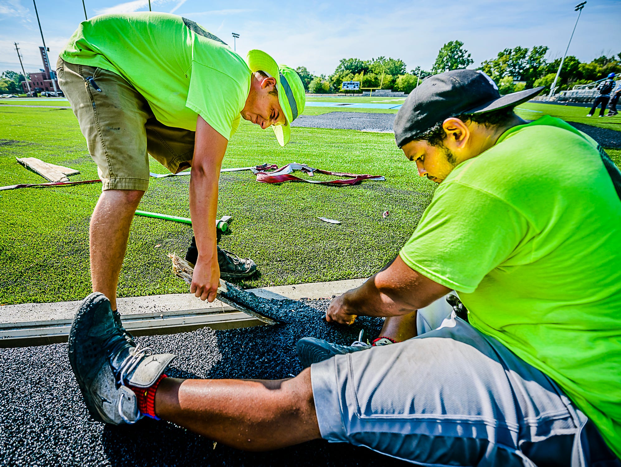 Michael Wheaton ,left, and Jeff Drye remove the extra wood that covers the drains between the new track and the football field at Lansing Catholics' new stadium.
