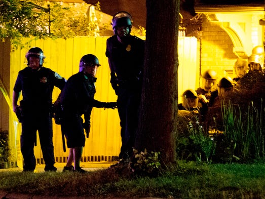 Police in riot gear crouch behind a tree and home after