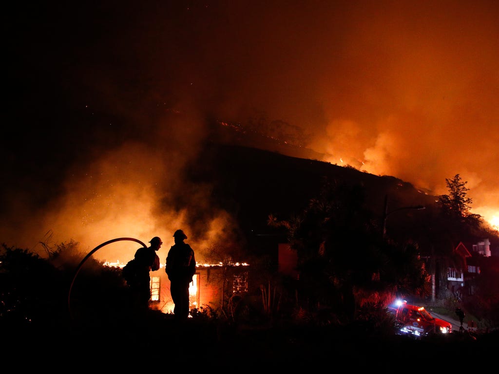 Two firefighters watch as a home burns in a wildfire in La Conchita, Calif., Dec. 7, 2017. The wind-swept blazes have forced tens of thousands of evacuations and destroyed dozens of homes.