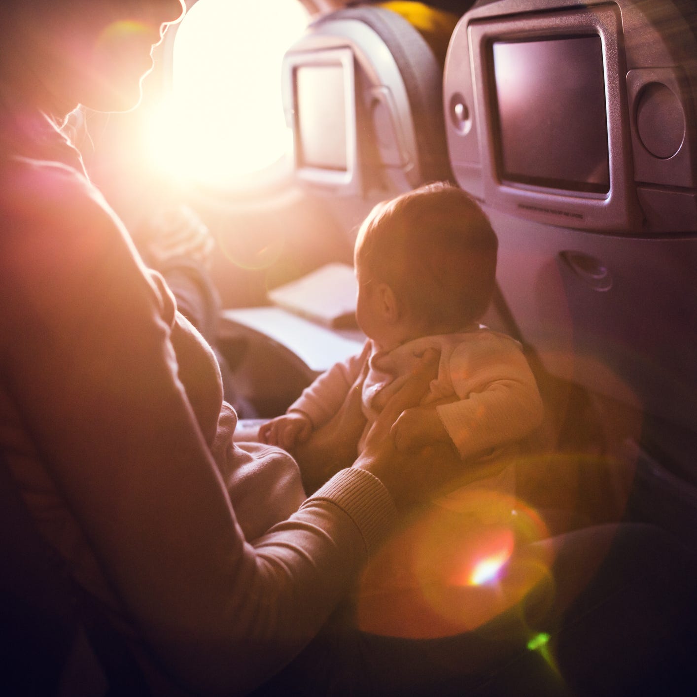 A mother and her baby girl sit in a passenger airline seat, the sun shining brightly in through the plane window.  While air travel with children can be difficult, both mom and child are content, the mother with a smile on her face. Horizontal image.  INTENTIONAL LENS FLARE.