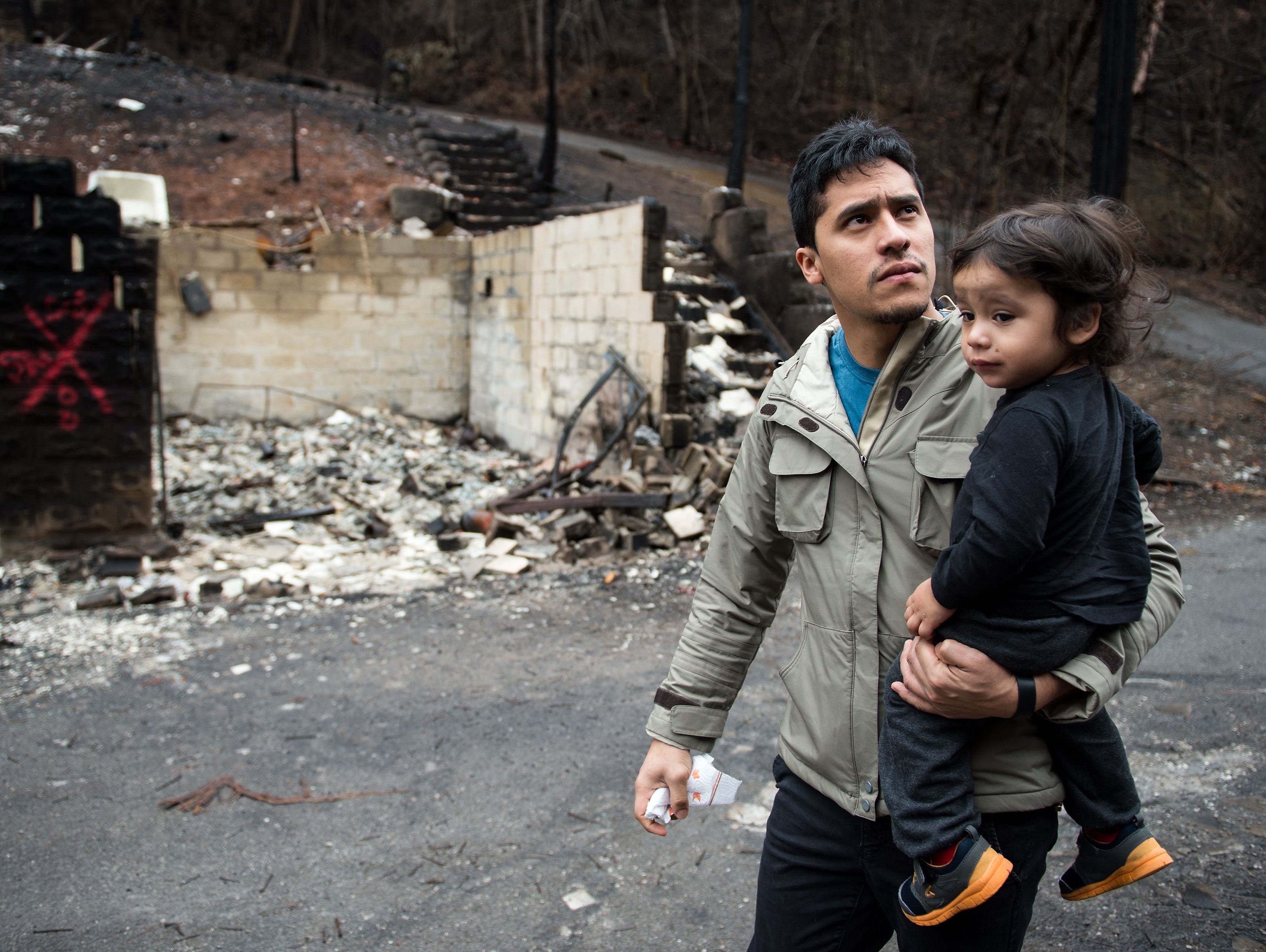 Allan Rivera holds onto his son Nathan Rivera, 23 months old, as he looks at the remains of their home for the first time. The family evacuated from their rental cabin before it was completely destroyed.