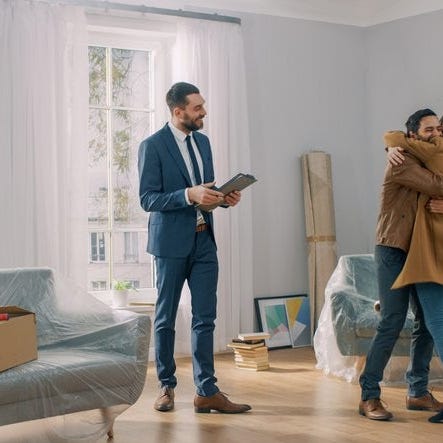 A man and woman hugging in the living room of their new house surrounded by moving boxes standing next to their realtor.