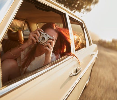 Woman taking a photo on vintage camera (pointed at viewer) on a rural road trip.