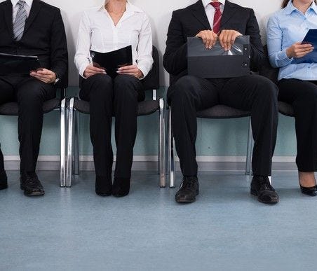 several people, shown from neck down, sitting on chairs next to each other, holding papers or materials that suggest they're waiting to be called in for an interview