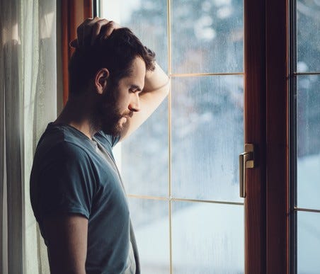 A man stands before a glass door, head on hand, head bowed, and eyes closed.