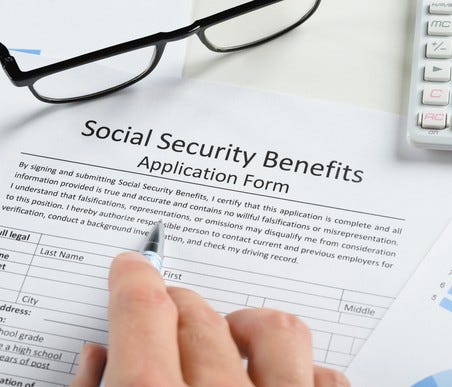 A hand filling out a Social Security benefit application form, next to a pair of reading glasses and a calculator