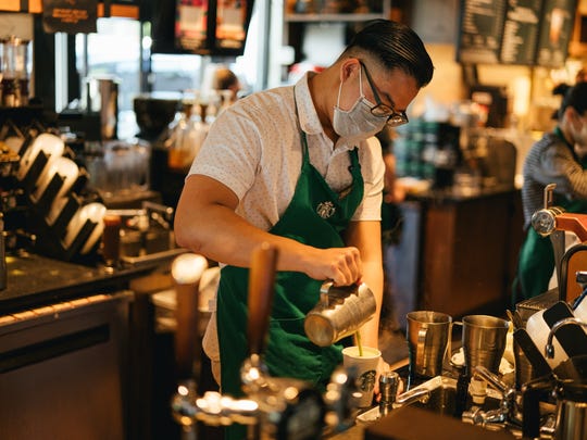 A Starbucks barista wears a mask and pours a coffee beverage.