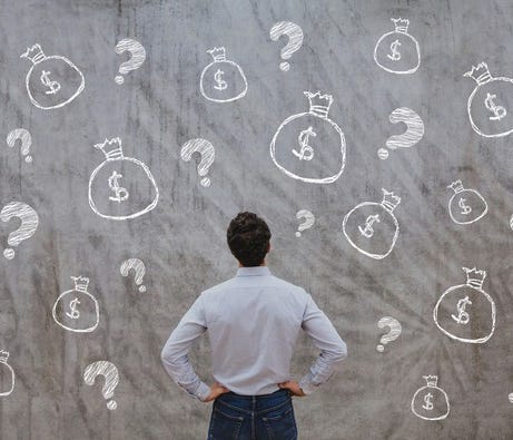 A man staring at a chalkboard with question marks and bags of money drawn on it.