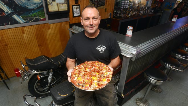 David Bailey, owner of Road Dogs Tavern on Ridge Avenue in Zanesville, holds one of the tavern's pizza while perched on one of the motorcyles that are seats at the bar.