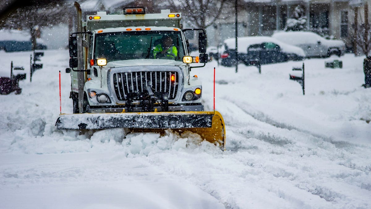 A work truck with yellow snow plow attachment clears a residential street after a winter storm.