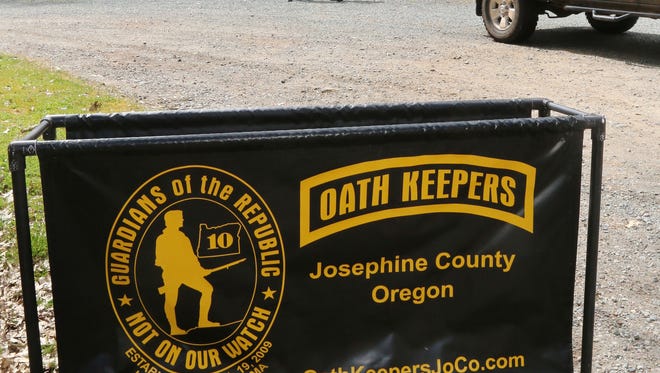In this photo taken Tuesday, April 14, 2015, an Oath Keepers, constitutional activists, sign marks the entrance to a property on Camp Joy Road near Merlin, Ore, while armed Oath Keepers security guards hover in the background. The site is a gathering of people who support the rights of miners on a claim near Galice, Oregon, that the Bureau of Land Management wants to bring into compliance or shut down. (Timothy Bullard/The Daily Courier via AP)