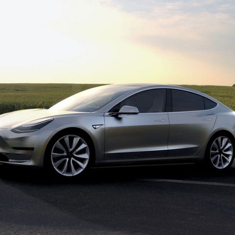 A silver Tesla Model 3 on a road, with a green fie