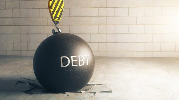A black wrecking ball, with the word DEBT written on it, on top of a floor that it has obviously broken upon landing