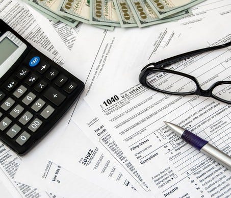 Tax forms, calculator, glasses, pen, and $100 bills laid on a surface