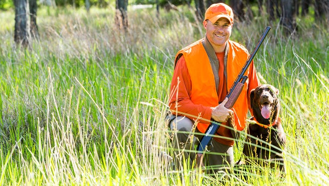 Hunter kneeling on the ground in tall grass, smiling at the camera holding a shotgun.  His hunting dog, a brown labrador retriever, is sitting next to him.  He is wearing an orange safety vest.