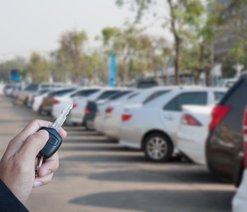 A hand holding a car key in the foreground, a row of vehicles at an auto dealership in the background.
