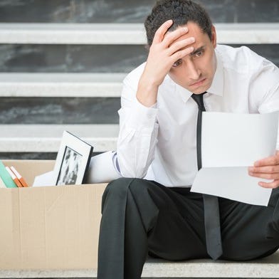 Man clutching his head while holding a piece of paper and sitting next to a box of desk supplies
