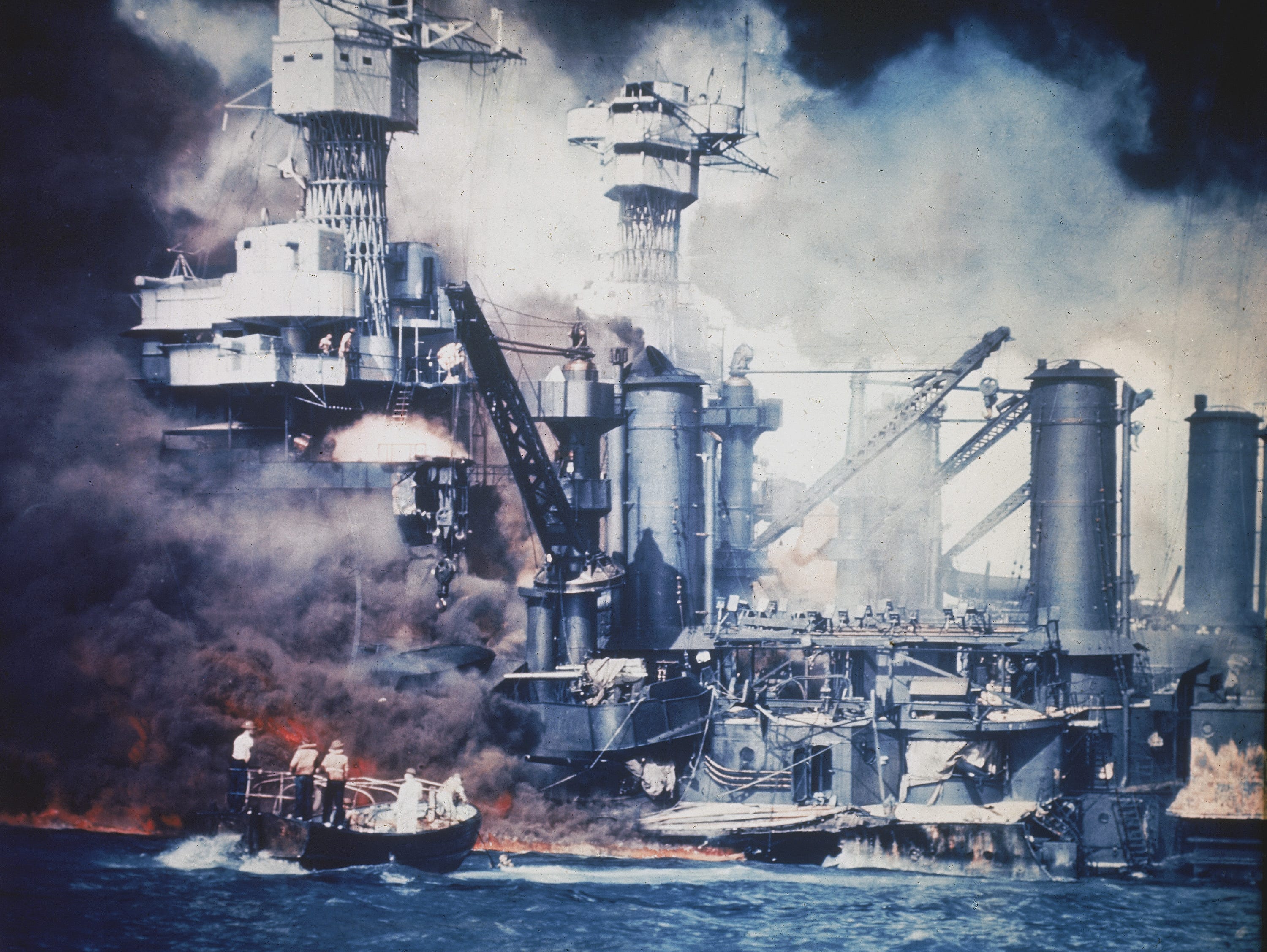 A small boat rescues a USS West Virginia crew members from the water after the Japanese bombing of Pearl Harbor. Two men can be seen on the superstructure, upper center. The mast of the USS Tennessee is beyond the burning West Virginia.