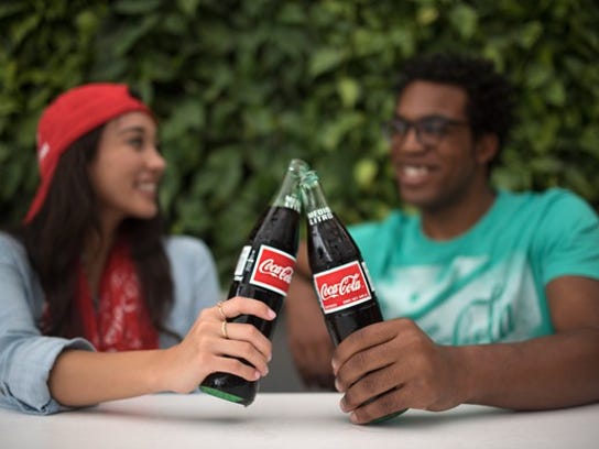 A young woman with a red hat and a young man with a green shirt are shown clinking their glass Coke bottles together