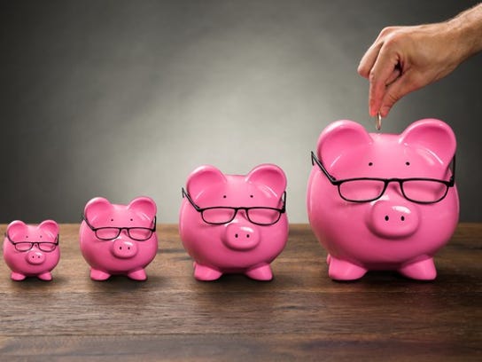 A series of progressively larger pink piggy banks wearing glasses, with a man's hand holding a coin over the largest one.