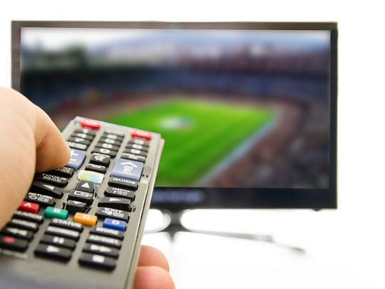 A TV broadcasting a sporting event is in the background. In the foreground is someone's hand holding a remote control pointed at the TV.