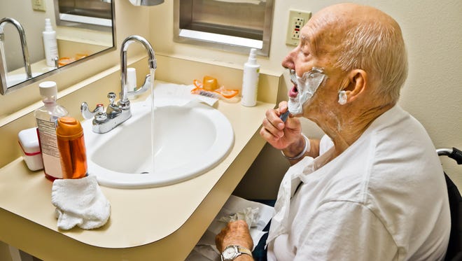 Elderly white male sitting in a wheelchair shaving at a handicap accommodating sink.
