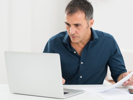 A man is sitting watching a laptop monitor while holding a document in his hand.