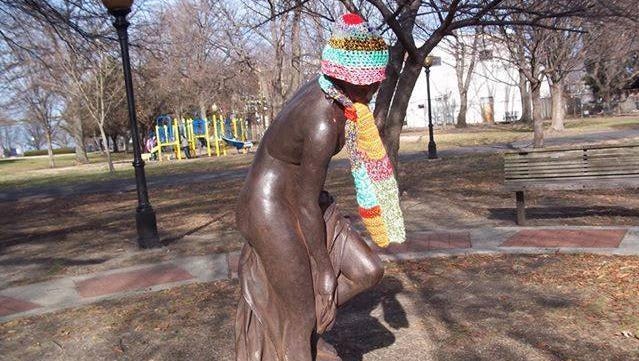 Colorful crochet hat and scarves are placed on Sadie, a statue in Proprietor's Park in Gloucester. A local woman makes the hats, dresses Sadie, and hopes the needy will take the items.