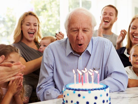Older person blowing out candles on cake while family members clap and look on in the background.