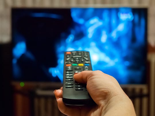 A hand holds a TV remote pointed at a big-screen television set in the background.