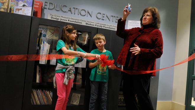 Kaye Price-Hawkins lifts a small pair of craft scissors, having discarded a ceremonially larger pair which failed to cut the ribbon held by siblings Emily and Judson Mitchell, Wednesday at the dedication of a family reading area at The Grace Museum. Price-Hawkins donated a children's book collection to the museum.