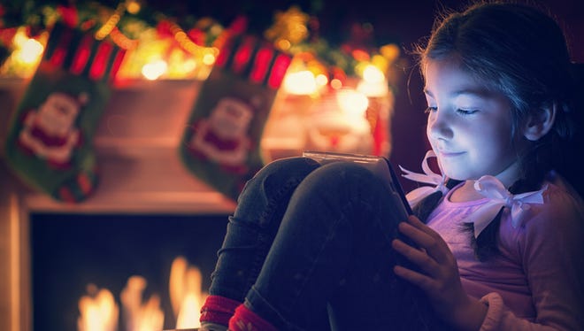 Smiling little girl sitting on the bed at night in front of fireplace at Christmas time. She enjoy while warming,playing and using digital tablet