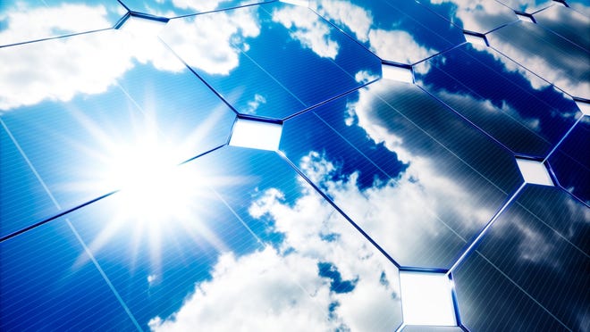 Close-up photo of a solar panel reflecting the sun and patchy clouds against a deep blue sky.
