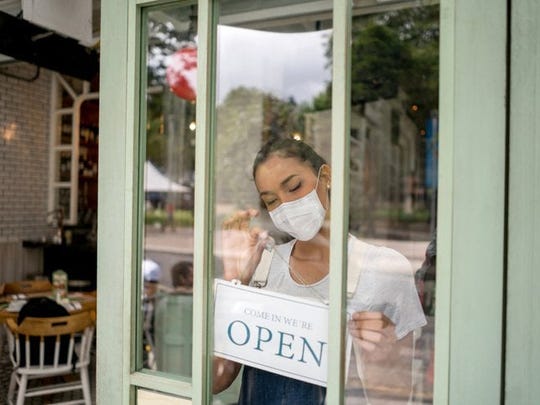 A small business owner wearing a mask and hanging an Open sign on the front door of her shop.
