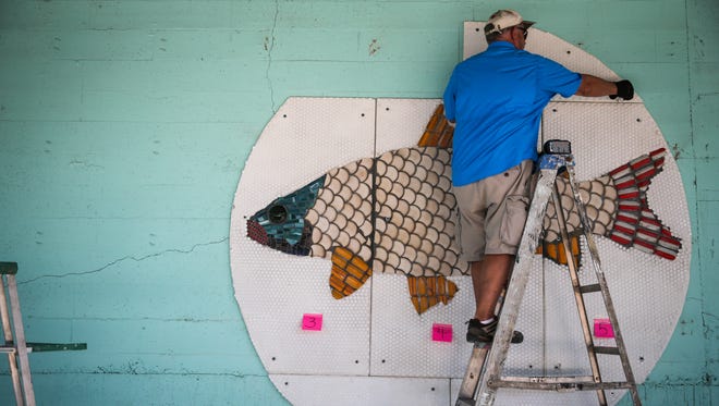 Jim Raymond helps with installation of a giant mosaic fish artwork Wednesday, June 14, on the wall facing The Bosque. 