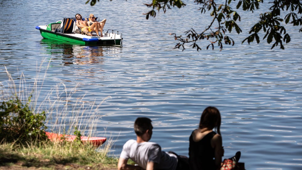 People sunbathe on a pedal boat as others rest in the shade of a tree on the bank of the Spree river, in Berlin, Germany.