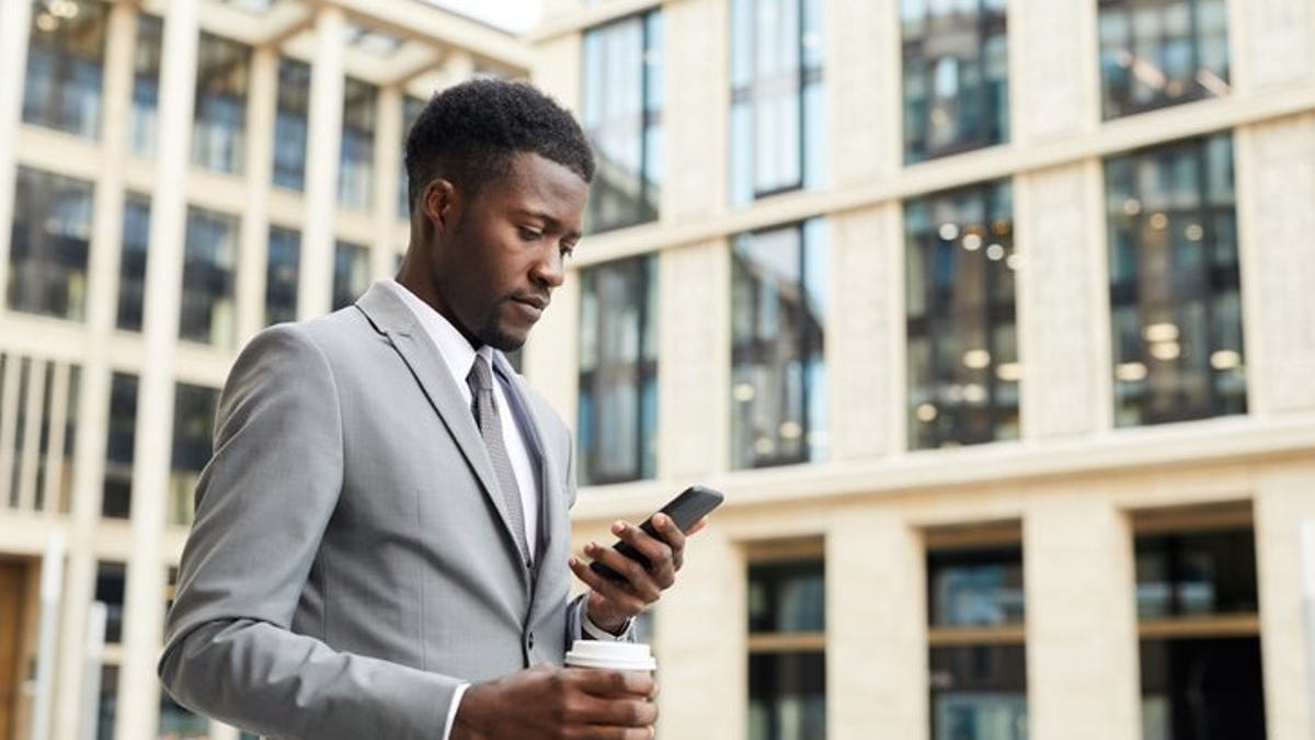 A man walking outside an office building while holding a coffee and looking at his phone.