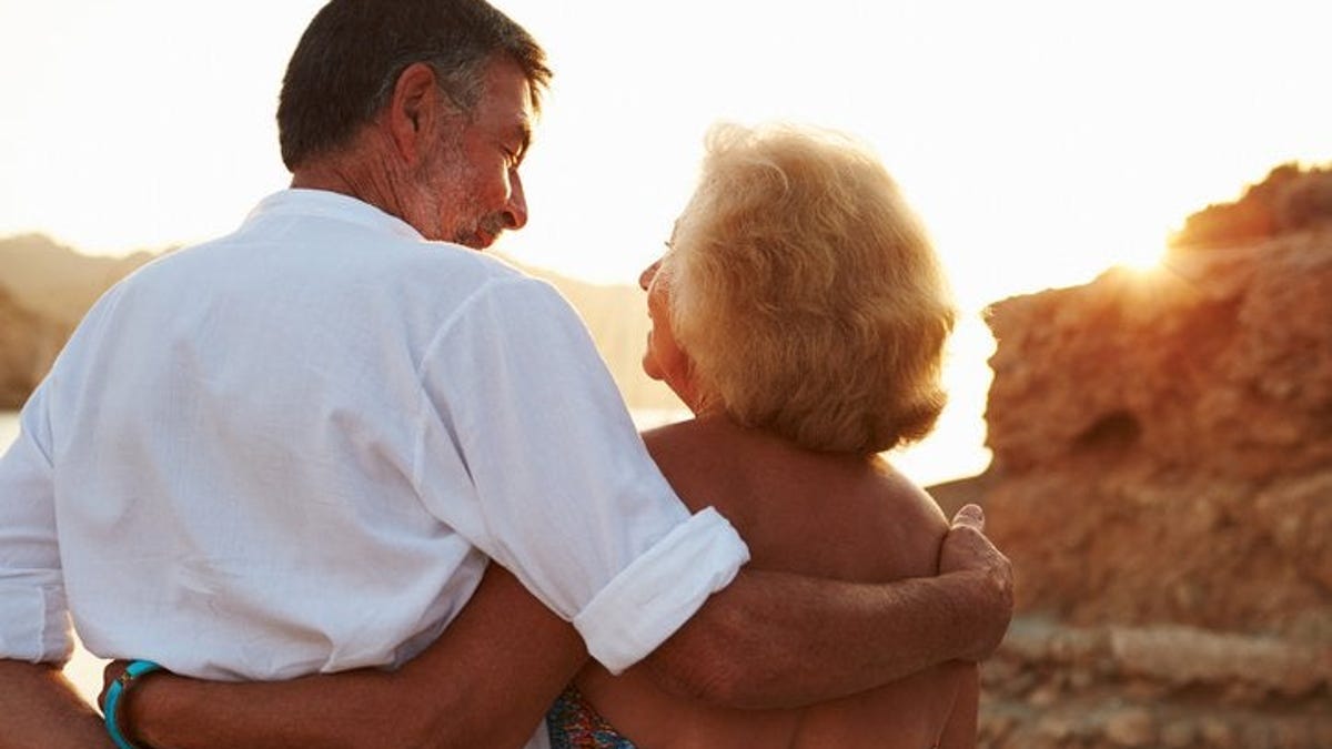 An older couple standing on the beach and looking at the sunset with their arms around each other.