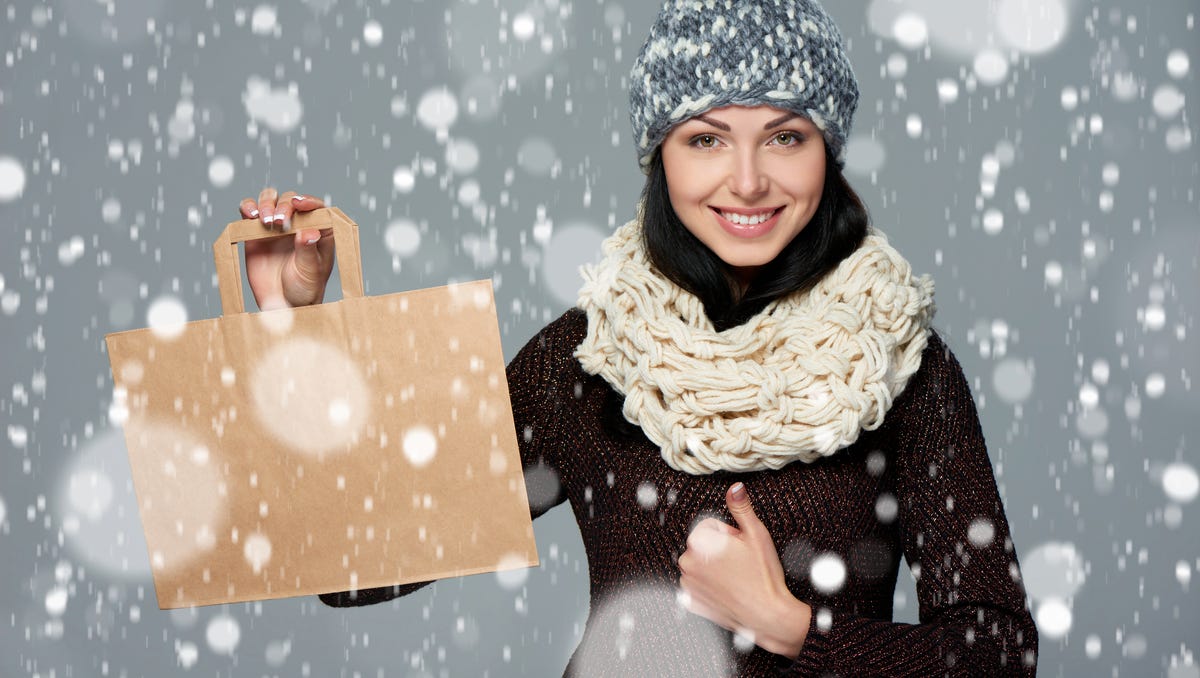 Winter holidays sale, shopping, Christmas concept. Portrait of smiling woman wearing warm winter hat and muffler showing shopping bag with empty copy space and gesturing thumb up over snow background