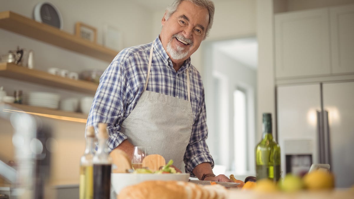 Smiling senior man wearing an apron and standing in front of a kitchen counter with olive oil, bread, and a salad on top of it.