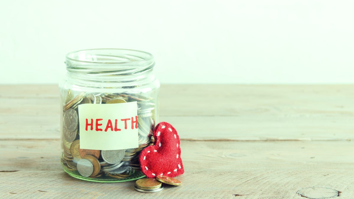 Coin-filled glass jar, sitting on wooden surface, labeled HEALTH, with red heart resting against it.