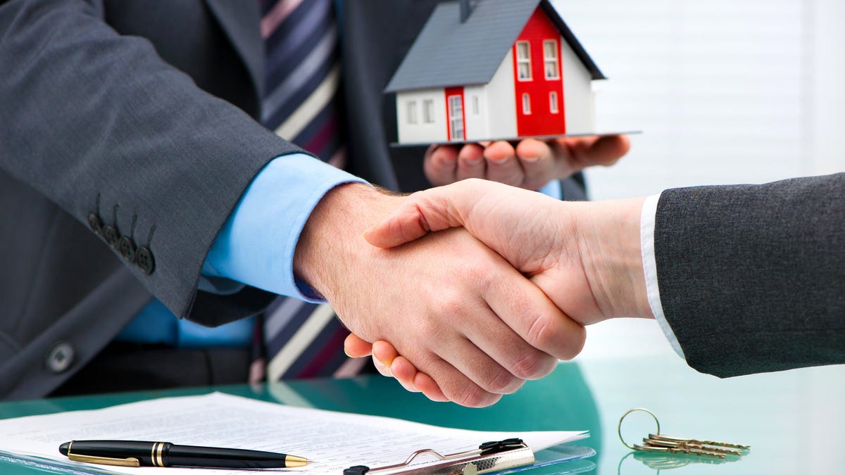 Two businesspersons shaking hands, with one holding a miniature house in his left hand.