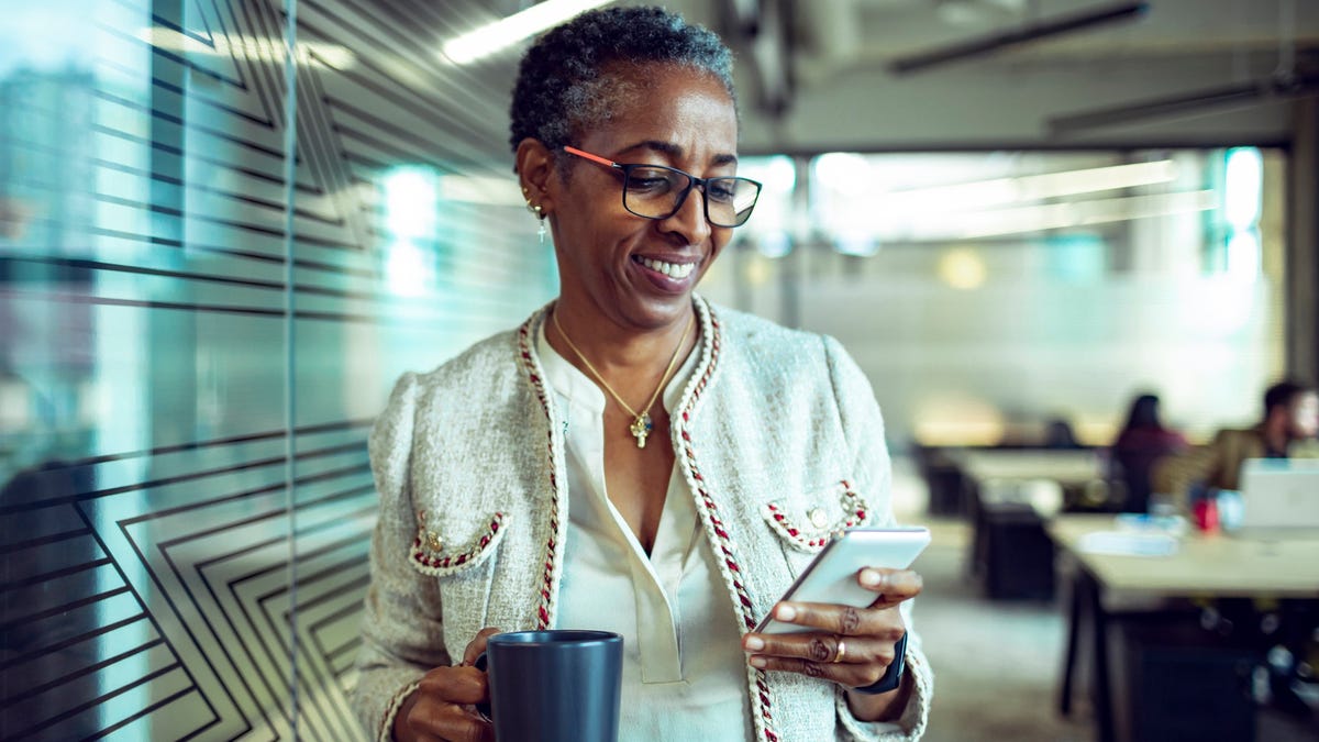 Person in office hallway, holding coffee cup and looking at smartphone.