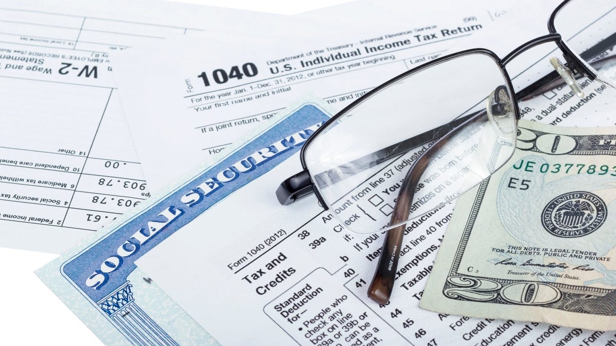 A Social Security card wedged between U.S. tax forms, with eyeglasses and a twenty-dollar bill set atop the tax forms.