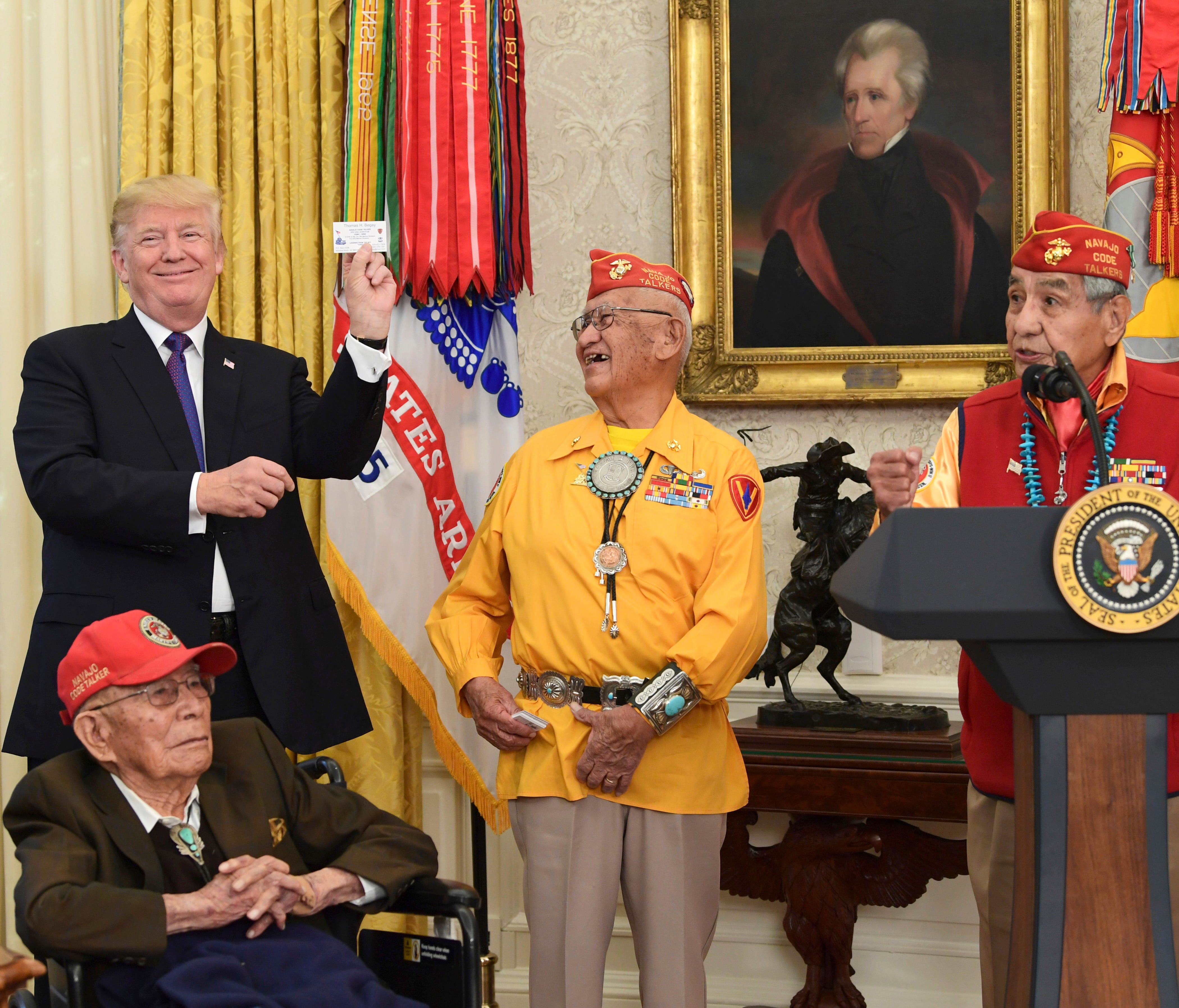 President Donald Trump, standing left, holds up the card of Navajo Code Talker Thomas Begay, center, during their meeting in the Oval Office of the White House in Washington, Monday, Nov. 27, 2017.