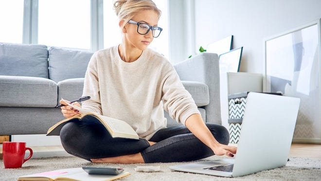 A young woman sitting on the floor of her living room with a notebook, calculator, and laptop.