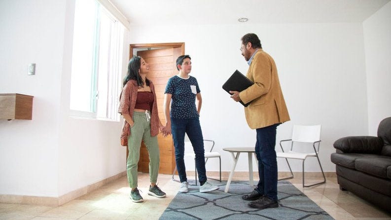 Two people holding hands while standing in the living room of a new house and talking with a realtor.