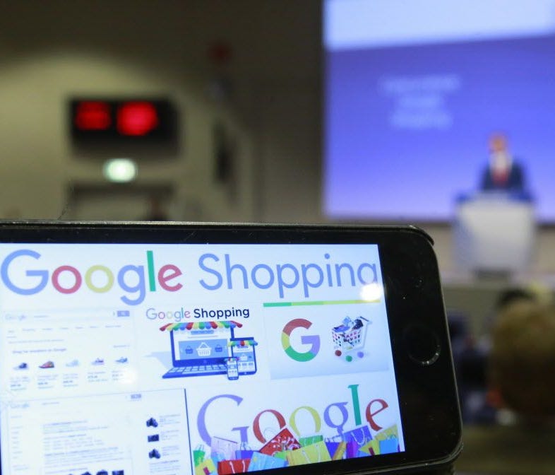Google Shopping logos on a phone as European Commissioner for Competition Margrethe Vestager (R) speaks during a press conference on an antitrust case against Google Shopping.
