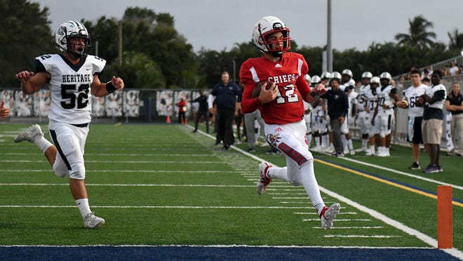 Quarterback Brody Palhegyi (12) of Cardinal Gibbons scores a touchdown in the first quarter as Ruben Navarro (52) of American Heritage follows on the play. Cardinal Gibbons vs. American Heritage School. Delray Beach, September 20, 2019.